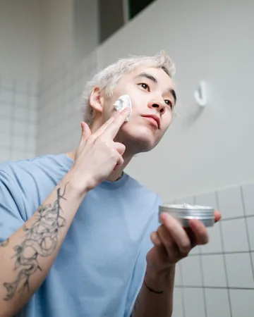 Uploaded photo, portrait: a man with bleached hair rubbing cream on his cheek, a tin in his other hand.