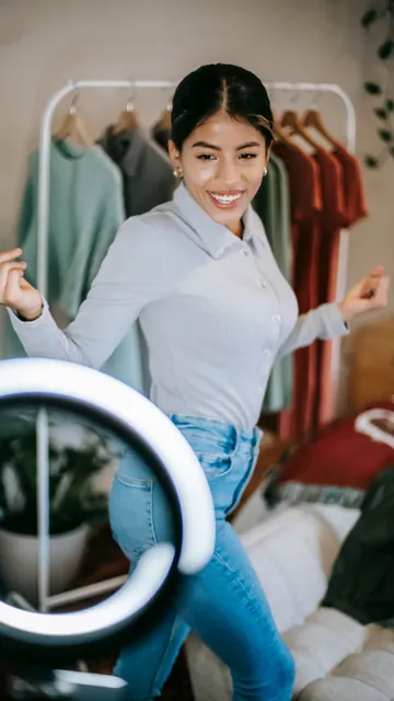 Uploaded photo, vertical: a woman showing an outfit to a phone on a ring light, a clothes rail behind her.